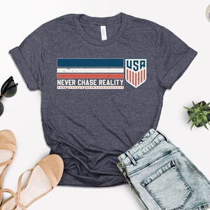 May include: Dark navy blue t-shirt with the text "NEVER CHASE REALITY" and a graphic of the USA flag. The shirt has a crew neck and short sleeves. Sunglasses and sandals are next to the shirt.