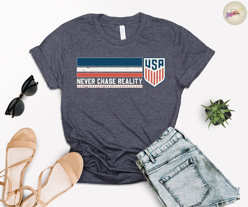 May include: Dark navy blue t-shirt with the text "NEVER CHASE REALITY" and a graphic of the USA flag. The shirt has a crew neck and short sleeves. Sunglasses and sandals are next to the shirt.