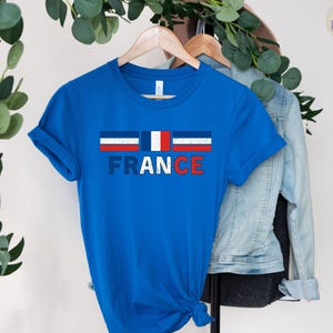 May include: A blue t-shirt with the word "FRANCE" in white and red letters. Above the word is a graphic of the French flag. The shirt is hanging on a wooden hanger.