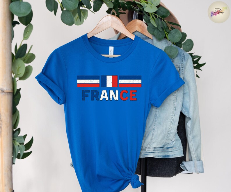 May include: A blue t-shirt with the word "FRANCE" in white and red letters. Above the word is a graphic of the French flag. The shirt is hanging on a wooden hanger.