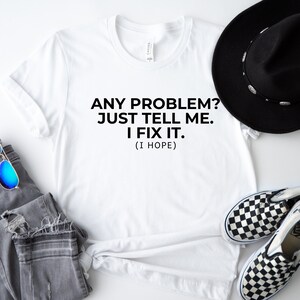 May include: A white t-shirt with the text "ANY PROBLEM? JUST TELL ME. I FIX IT. (I HOPE)". The shirt is paired with grey jeans, blue sunglasses, a black hat, and checkered slip-on shoes.