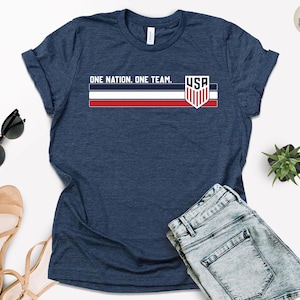 Może przedstawiać: Granatowa koszulka z napisem "ONE NATION. ONE TEAM." i grafiką flagi USA. Koszulka ma okrągły dekolt i kr&oacute;tkie rękawy. Na zdjęciu widoczne są r&oacute;wnież dżinsowe szorty, okulary przeciwsłoneczne i sandały.