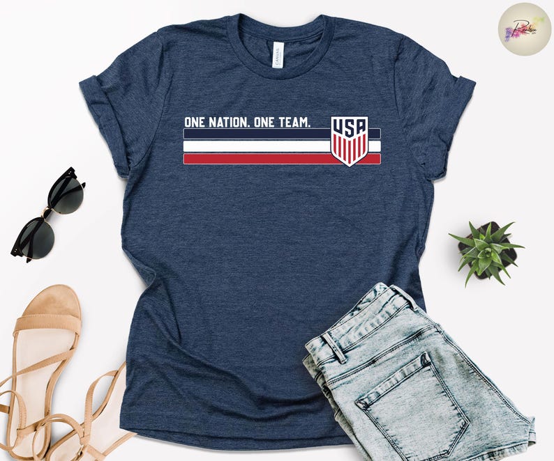 Może przedstawiać: Granatowa koszulka z napisem "ONE NATION. ONE TEAM." i grafiką flagi USA. Koszulka ma okrągły dekolt i kr&oacute;tkie rękawy. Na zdjęciu widoczne są r&oacute;wnież dżinsowe szorty, okulary przeciwsłoneczne i sandały.