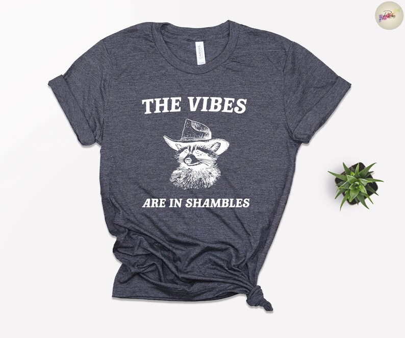 K&ouml;nnte beinhalten: Ein dunkel-helles graues T-Shirt mit einer wei&szlig;en Grafik eines Waschb&auml;ren, der einen Cowboyhut tr&auml;gt. Der Text "THE VIBES ARE IN SHAMBLES" ist auf das T-Shirt gedruckt.