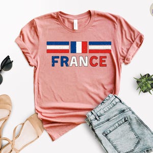 May include: A light pink t-shirt with the word "FRANCE" in red, white, and blue letters. Above the word is a graphic of the French flag. The shirt is displayed with sunglasses, sandals, and denim shorts.