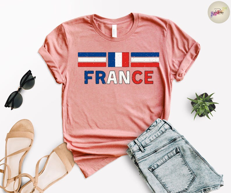 May include: A light pink t-shirt with the word "FRANCE" in red, white, and blue letters. Above the word is a graphic of the French flag. The shirt is displayed with sunglasses, sandals, and denim shorts.