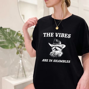 K&ouml;nnte beinhalten: Schwarzes T-Shirt mit einer wei&szlig;en Grafik eines Waschb&auml;ren, der einen Cowboyhut tr&auml;gt. Der Text "THE VIBES ARE IN SHAMBLES" ist unter dem Waschb&auml;ren gedruckt.