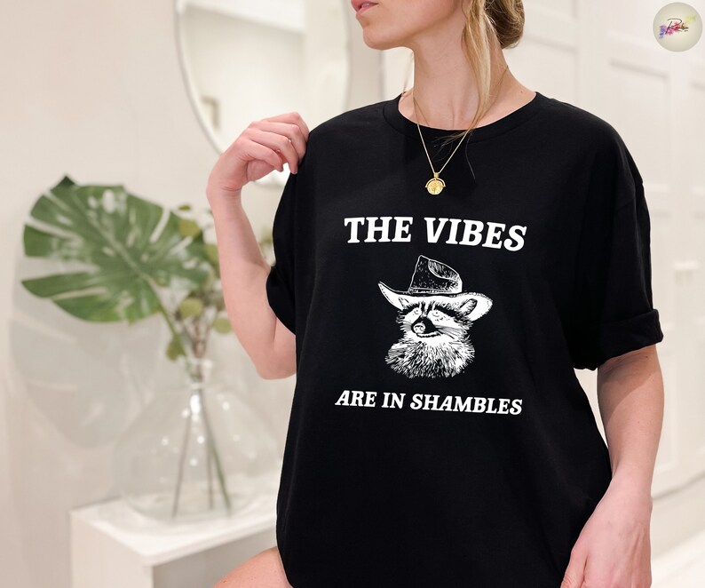 K&ouml;nnte beinhalten: Schwarzes T-Shirt mit einer wei&szlig;en Grafik eines Waschb&auml;ren, der einen Cowboyhut tr&auml;gt. Der Text "THE VIBES ARE IN SHAMBLES" ist unter dem Waschb&auml;ren gedruckt.