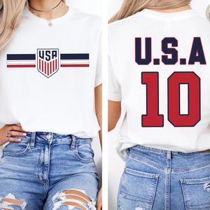 May include: A white short-sleeve t-shirt featuring a USA shield logo with horizontal navy and red stripes on the front. The back of the shirt displays the text U.S.A. in navy blue above the large red number 10 with a navy blue outline.