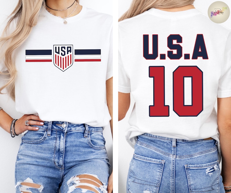 May include: A white short-sleeve t-shirt featuring a USA shield logo with horizontal navy and red stripes on the front. The back of the shirt displays the text U.S.A. in navy blue above the large red number 10 with a navy blue outline.