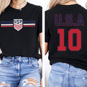 May include: A black short-sleeve t-shirt featuring a front graphic of a shield with the text USA and horizontal stripes in navy, white, and red. The back of the shirt displays the text U.S.A. in navy blue above the large red number 10.