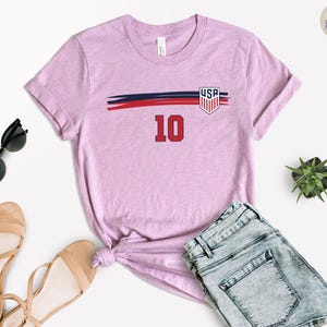 May include: A light purple short-sleeve t-shirt featuring a horizontal navy blue and red brushstroke graphic across the chest. The design includes a shield emblem with the text USA and the number 10 in red with a black outline centered below.