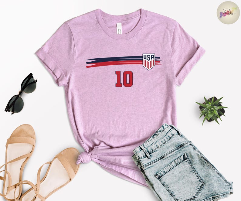 May include: A light purple short-sleeve t-shirt featuring a horizontal navy blue and red brushstroke graphic across the chest. The design includes a shield emblem with the text USA and the number 10 in red with a black outline centered below.