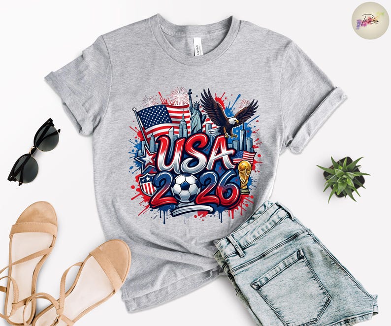 May include: A light gray t-shirt with a colorful graphic design featuring the text "USA 2026", an American flag, a soccer ball, and an eagle. The design also includes the Statue of Liberty and a gold trophy.