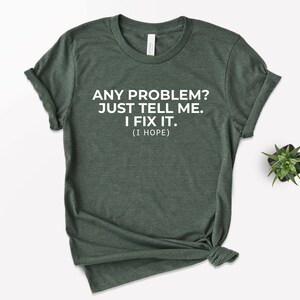 May include: Dark green t-shirt with white text: "ANY PROBLEM? JUST TELL ME. I FIX IT. (I HOPE)." The shirt is knotted at the bottom. A small succulent plant is visible in the background.