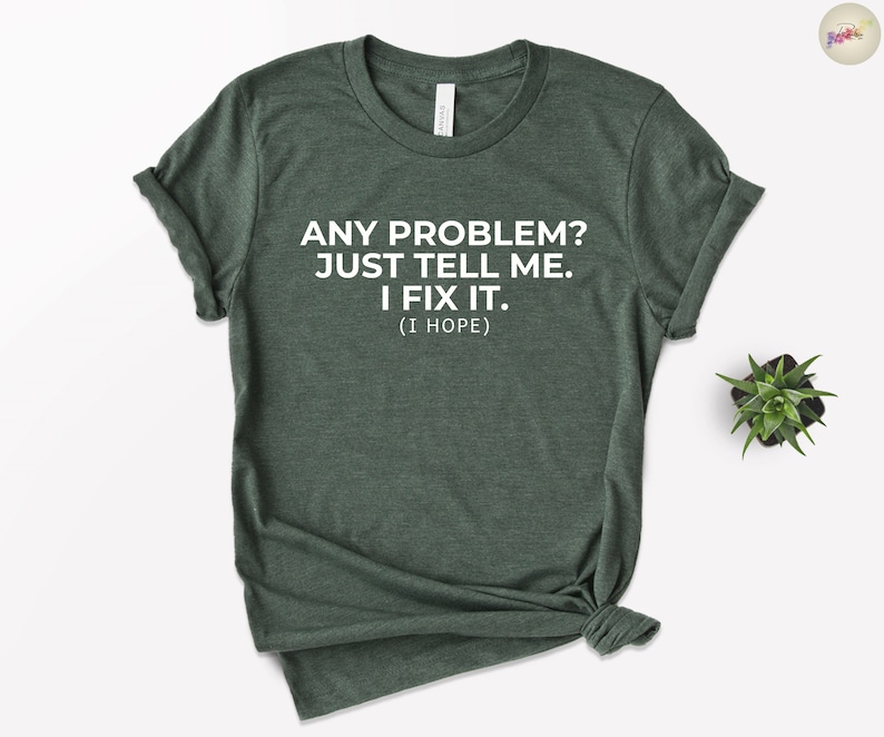 May include: Dark green t-shirt with white text: "ANY PROBLEM? JUST TELL ME. I FIX IT. (I HOPE)." The shirt is knotted at the bottom. A small succulent plant is visible in the background.