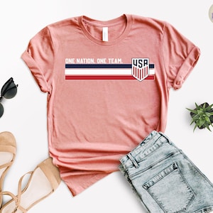 Może przedstawiać: Koralowa koszulka z napisem "ONE NATION. ONE TEAM." i logo USA. Projekt zawiera poziomy pasek w kolorze granatowym, białym i czerwonym. Koszulka jest prezentowana z okularami przeciwsłonecznymi, sandałami i szortami dżinsowymi.