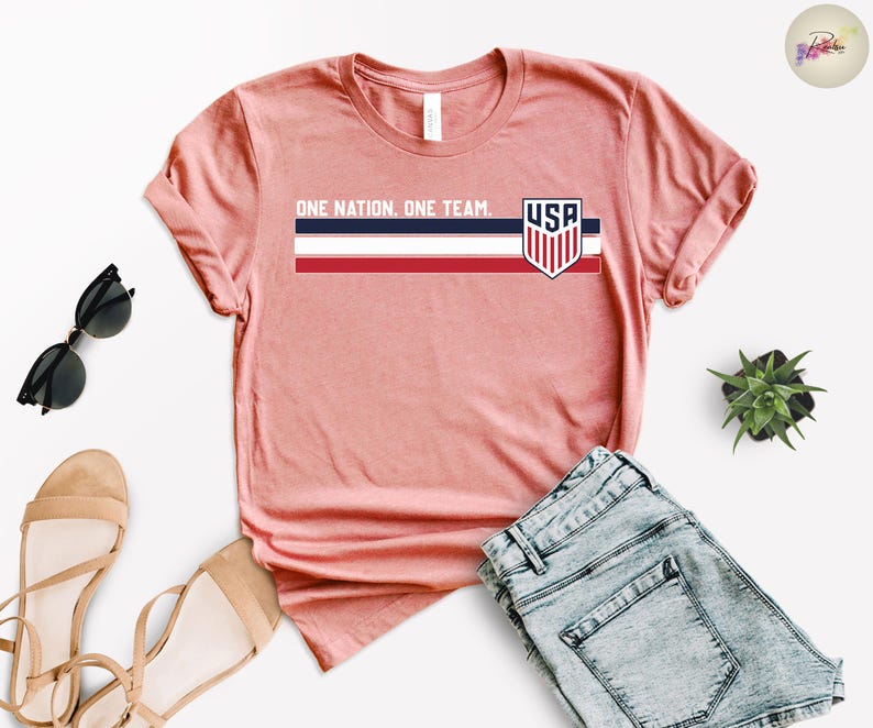 Może przedstawiać: Koralowa koszulka z napisem "ONE NATION. ONE TEAM." i logo USA. Projekt zawiera poziomy pasek w kolorze granatowym, białym i czerwonym. Koszulka jest prezentowana z okularami przeciwsłonecznymi, sandałami i szortami dżinsowymi.