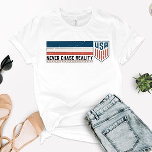 May include: White t-shirt with a graphic design featuring the text "NEVER CHASE REALITY" and a USA shield emblem. The design incorporates blue, red, and white colors. The image also includes sunglasses, sandals, and denim shorts.