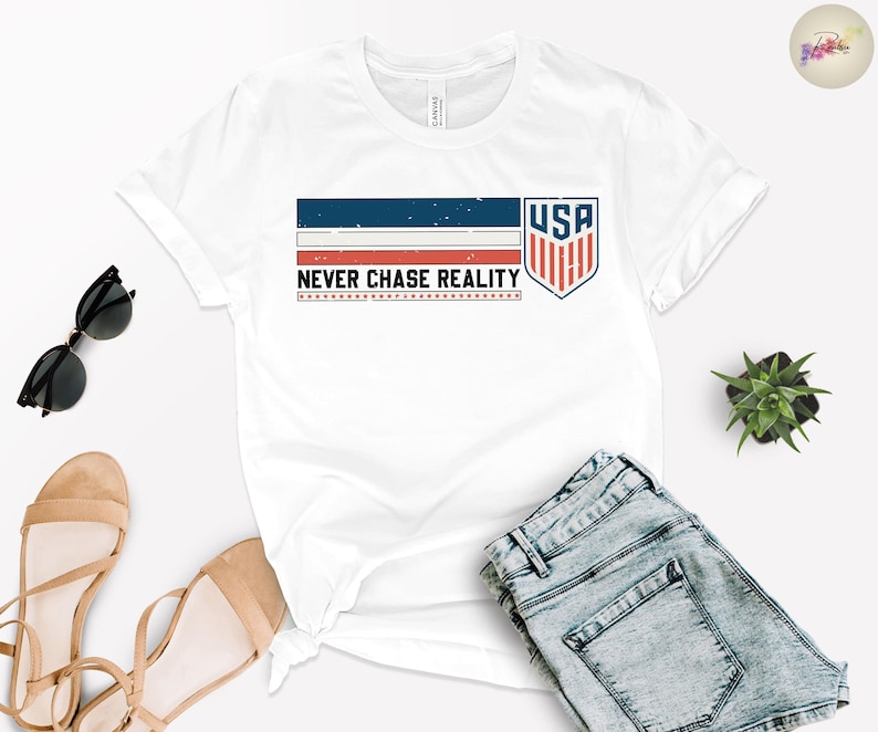 May include: White t-shirt with a graphic design featuring the text "NEVER CHASE REALITY" and a USA shield emblem. The design incorporates blue, red, and white colors. The image also includes sunglasses, sandals, and denim shorts.