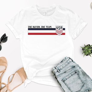 Może przedstawiać: Biała koszulka z napisem "ONE NATION. ONE TEAM." i emblematem USA w kolorach czerwonym, białym i niebieskim. Koszulka jest zestawiona z niebieskimi szortami dżinsowymi, okularami przeciwsłonecznymi i beżowymi sandałami. Widoczna jest r&oacute;wnież mała roślina.