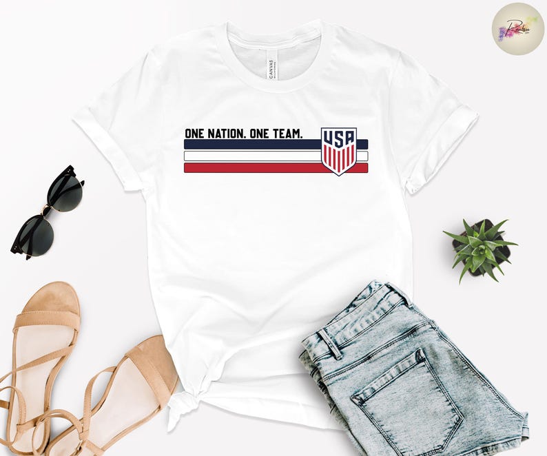 Może przedstawiać: Biała koszulka z napisem "ONE NATION. ONE TEAM." i emblematem USA w kolorach czerwonym, białym i niebieskim. Koszulka jest zestawiona z niebieskimi szortami dżinsowymi, okularami przeciwsłonecznymi i beżowymi sandałami. Widoczna jest r&oacute;wnież mała roślina.