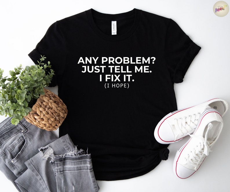 May include: Black t-shirt with the text "ANY PROBLEM? JUST TELL ME. I FIX IT. (I HOPE)". The shirt is paired with grey jeans, a woven basket, and white trainers with red accents.