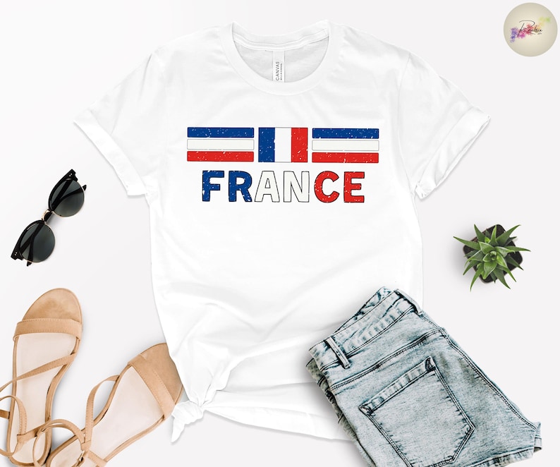 May include: White t-shirt with the word "FRANCE" in red, white, and blue, with a distressed flag design above the text. The shirt is paired with denim shorts, sunglasses, and sandals.
