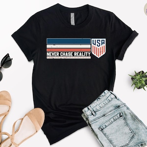 May include: Black t-shirt with a graphic design featuring the text "NEVER CHASE REALITY" and a USA shield emblem. The design incorporates red, white, and blue stripes. The shirt is paired with denim shorts and sandals.