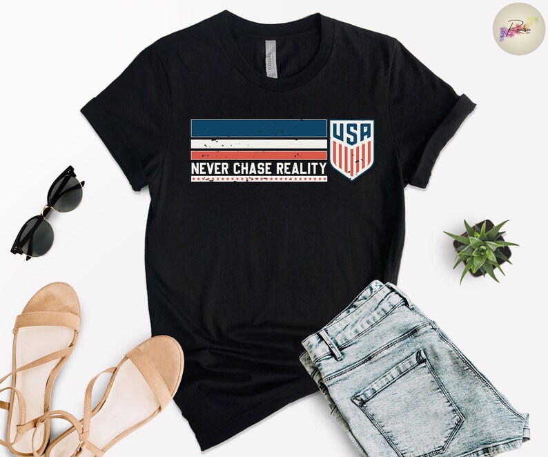 May include: Black t-shirt with a graphic design featuring the text "NEVER CHASE REALITY" and a USA shield emblem. The design incorporates red, white, and blue stripes. The shirt is paired with denim shorts and sandals.