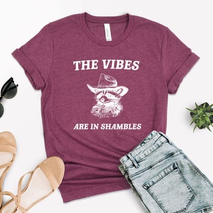 K&ouml;nnte beinhalten: Ein maroonfarbenes T-Shirt mit einer wei&szlig;en Grafik eines Waschb&auml;ren, der einen Cowboyhut tr&auml;gt. Der Text "THE VIBES ARE IN SHAMBLES" ist auf das Shirt gedruckt.