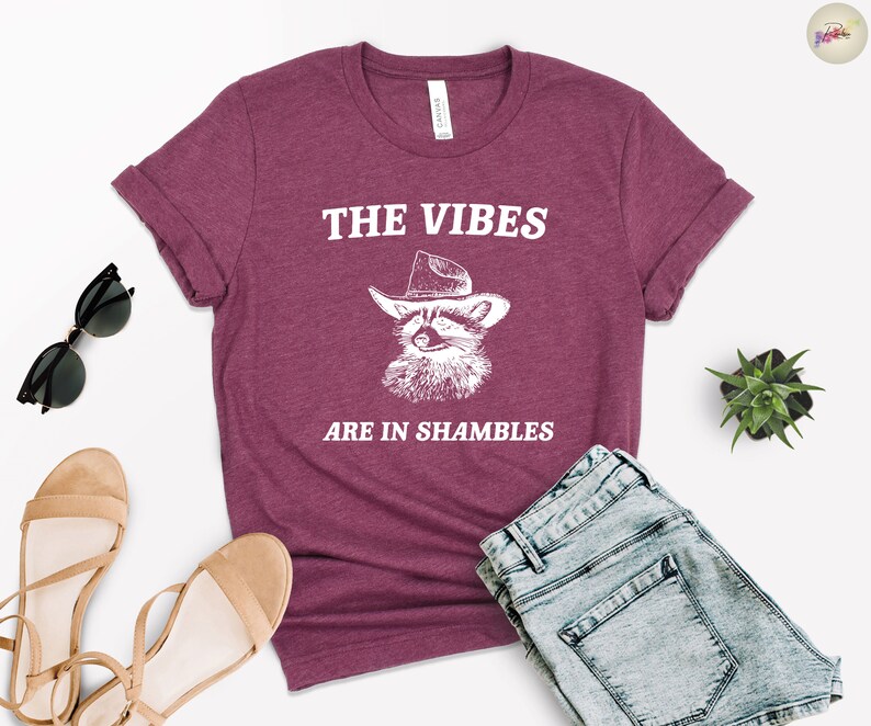 K&ouml;nnte beinhalten: Ein maroonfarbenes T-Shirt mit einer wei&szlig;en Grafik eines Waschb&auml;ren, der einen Cowboyhut tr&auml;gt. Der Text "THE VIBES ARE IN SHAMBLES" ist auf das Shirt gedruckt.