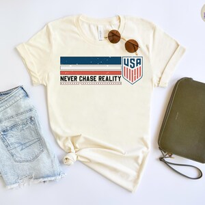 May include: Cream-colored t-shirt with a patriotic design featuring the text "USA" inside a shield, and the phrase "NEVER CHASE REALITY". The shirt is paired with denim shorts, sunglasses, and a green clutch.