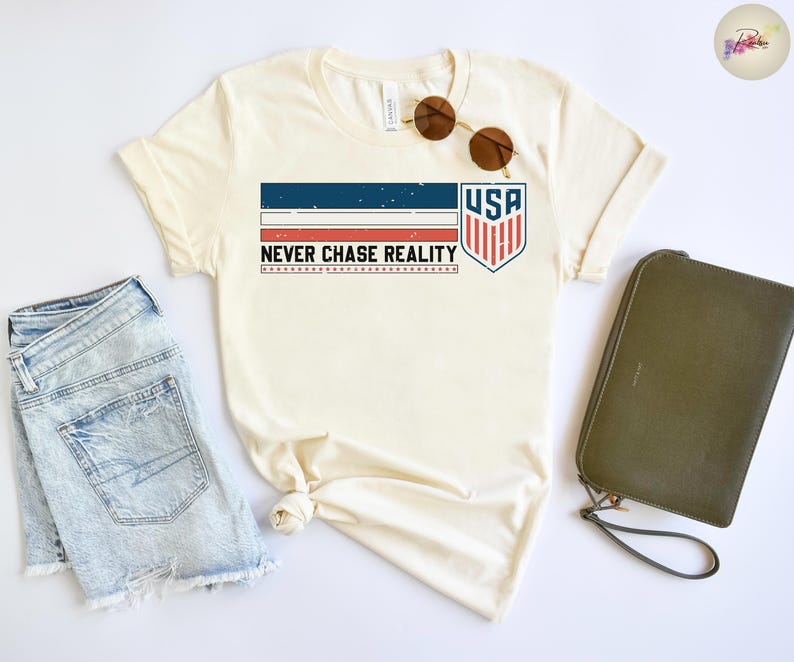 May include: Cream-colored t-shirt with a patriotic design featuring the text "USA" inside a shield, and the phrase "NEVER CHASE REALITY". The shirt is paired with denim shorts, sunglasses, and a green clutch.