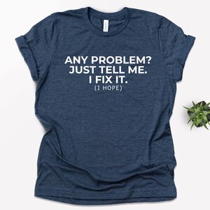 May include: A navy blue t-shirt with the text "ANY PROBLEM? JUST TELL ME. I FIX IT. (I HOPE)" printed in white. The shirt is a crew neck style and has rolled-up sleeves. A small green plant is visible in the background.
