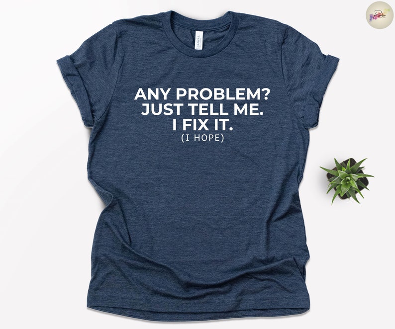 May include: A navy blue t-shirt with the text "ANY PROBLEM? JUST TELL ME. I FIX IT. (I HOPE)" printed in white. The shirt is a crew neck style and has rolled-up sleeves. A small green plant is visible in the background.