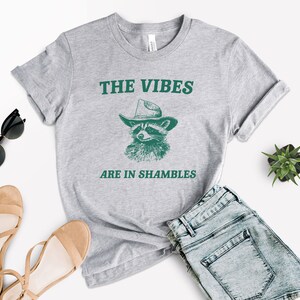 K&ouml;nnte beinhalten: Graues T-Shirt mit einer gr&uuml;nen Grafik eines Waschb&auml;ren, der einen Cowboyhut tr&auml;gt. Der Text "THE VIBES ARE IN SHAMBLES" ist auf das T-Shirt gedruckt.