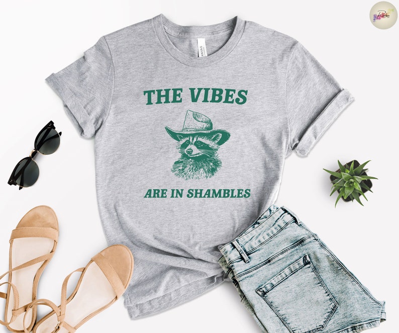 K&ouml;nnte beinhalten: Graues T-Shirt mit einer gr&uuml;nen Grafik eines Waschb&auml;ren, der einen Cowboyhut tr&auml;gt. Der Text "THE VIBES ARE IN SHAMBLES" ist auf das T-Shirt gedruckt.
