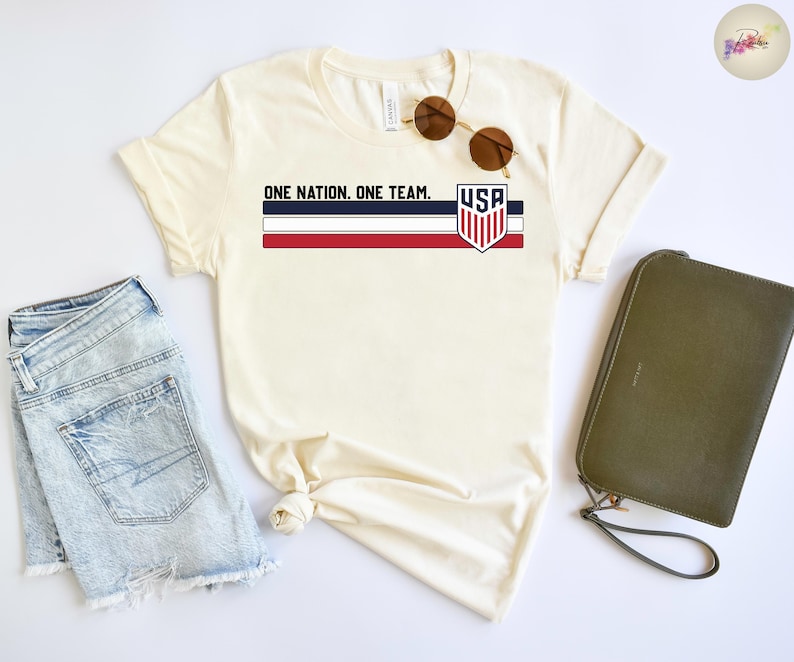 Może przedstawiać: Kremowa koszulka z napisem "ONE NATION. ONE TEAM." i logo tarczy USA. Koszulka ma czerwono-biało-niebieski wz&oacute;r w paski. Okulary przeciwsłoneczne i zielona kopert&oacute;wka leżą obok koszulki. Na zdjęciu są r&oacute;wnież jasnoniebieskie szorty dżinsowe.