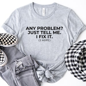 May include: A grey t-shirt with the text "ANY PROBLEM? JUST TELL ME. I FIX IT. (I HOPE)". The shirt is styled with grey jeans, black and white checkered slip-on shoes, and a black and white patterned hat.