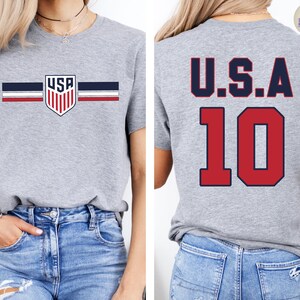May include: A heather gray short-sleeve t-shirt featuring a small USA shield logo with horizontal navy and red stripes on the front. The back of the shirt displays the text U.S.A. in navy blue above the large red number 10 with a navy blue outline.