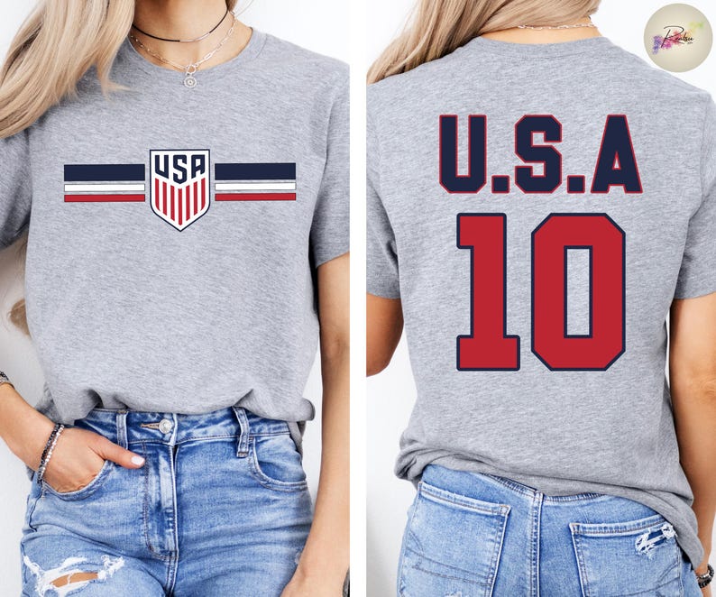May include: A heather gray short-sleeve t-shirt featuring a small USA shield logo with horizontal navy and red stripes on the front. The back of the shirt displays the text U.S.A. in navy blue above the large red number 10 with a navy blue outline.