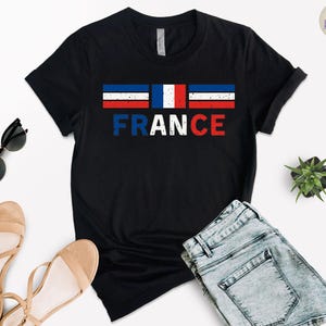 May include: Black t-shirt with the word "FRANCE" in white, distressed font. Above the word is a horizontal tricolor design in blue, white, and red, representing the French flag. The shirt is paired with denim shorts and sandals.