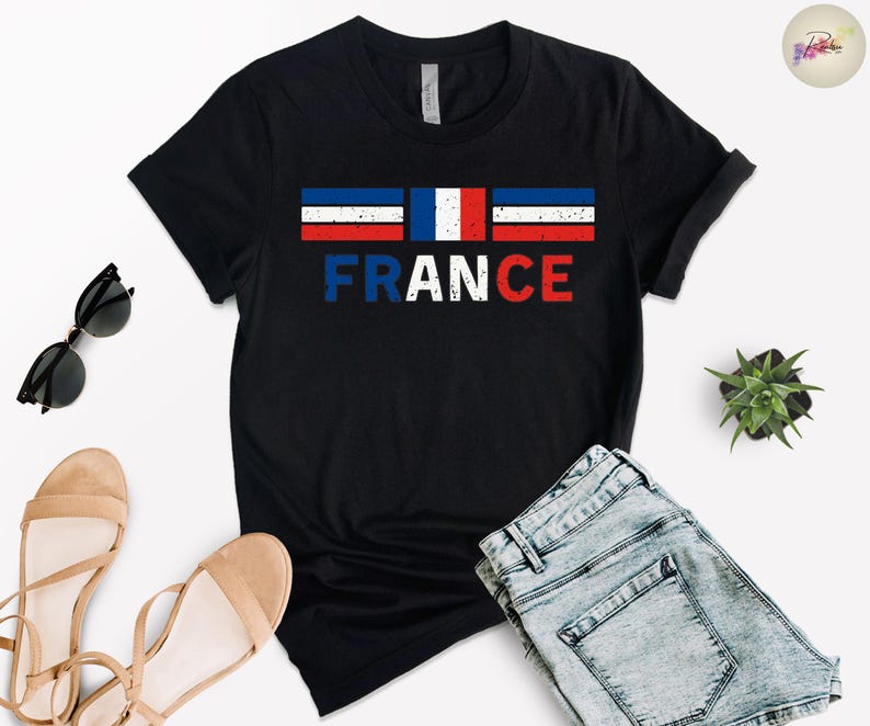 May include: Black t-shirt with the word "FRANCE" in white, distressed font. Above the word is a horizontal tricolor design in blue, white, and red, representing the French flag. The shirt is paired with denim shorts and sandals.