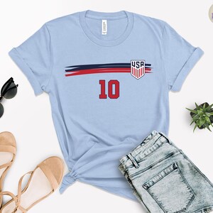 May include: A light blue short-sleeve t-shirt featuring a horizontal navy and red brushstroke graphic across the chest. The design includes a shield-shaped USA emblem and the number 10 in red with a black outline centered below the graphic.