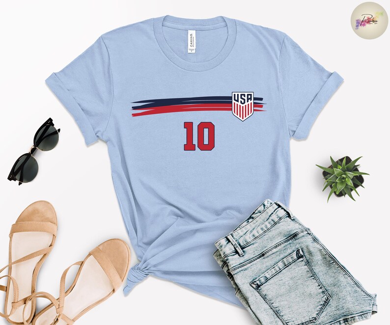 May include: A light blue short-sleeve t-shirt featuring a horizontal navy and red brushstroke graphic across the chest. The design includes a shield-shaped USA emblem and the number 10 in red with a black outline centered below the graphic.