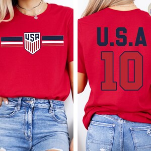 May include: A red short-sleeve t-shirt featuring a USA shield logo with horizontal stripes on the front and the text U.S.A. with the number 10 in large dark blue font on the back. The shirt is paired with blue denim jeans.
