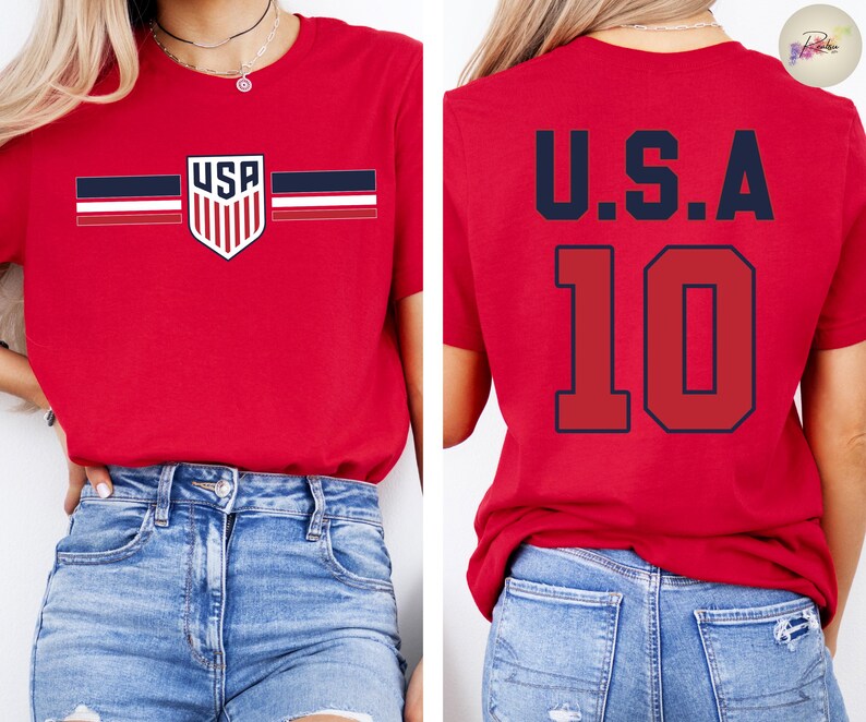 May include: A red short-sleeve t-shirt featuring a USA shield logo with horizontal stripes on the front and the text U.S.A. with the number 10 in large dark blue font on the back. The shirt is paired with blue denim jeans.