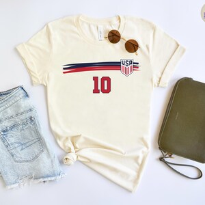 May include: A cream-colored short-sleeve t-shirt featuring a graphic with a horizontal navy and red brushstroke design, a USA shield emblem, and the number 10 in red. The shirt is displayed alongside light blue denim shorts and a dark olive clutch.