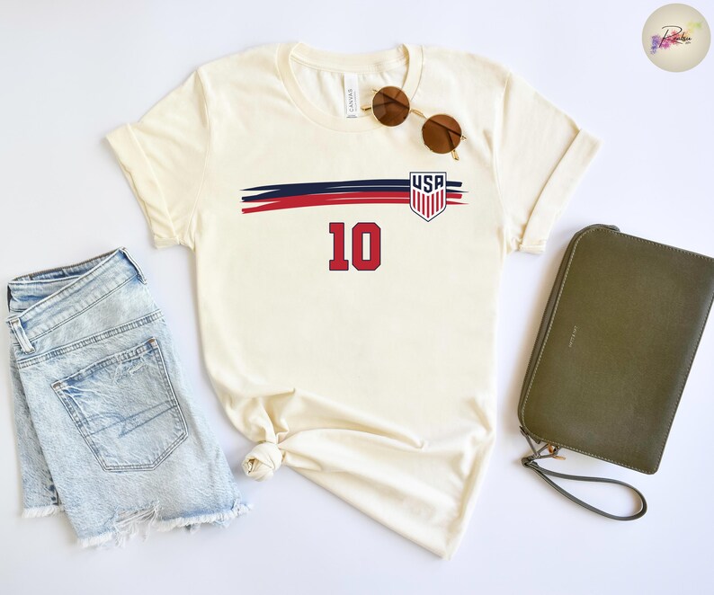 May include: A cream-colored short-sleeve t-shirt featuring a graphic with a horizontal navy and red brushstroke design, a USA shield emblem, and the number 10 in red. The shirt is displayed alongside light blue denim shorts and a dark olive clutch.